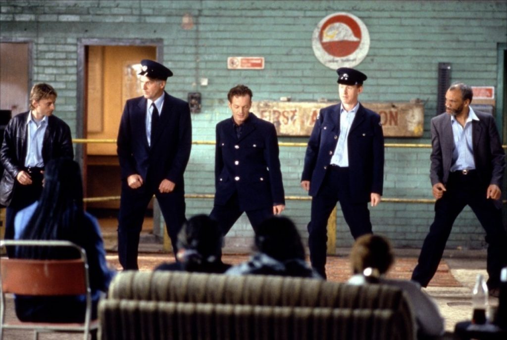 Seis hombres en trajes formales de pie en un interior industrial, con sofá desgastado al frente