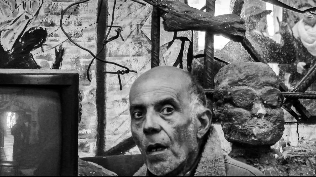 Documental "La memoria al revés": El dilema de las apariencias Hombre mayor hablando con expresión seria junto a una escultura de rostro, en blanco y negro, escena de documental