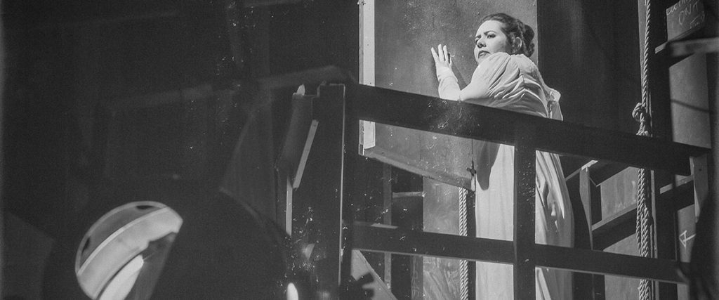 Escena de teatro en blanco y negro: actor en plataforma elevada observa a otro personaje abajo, iluminación dramática