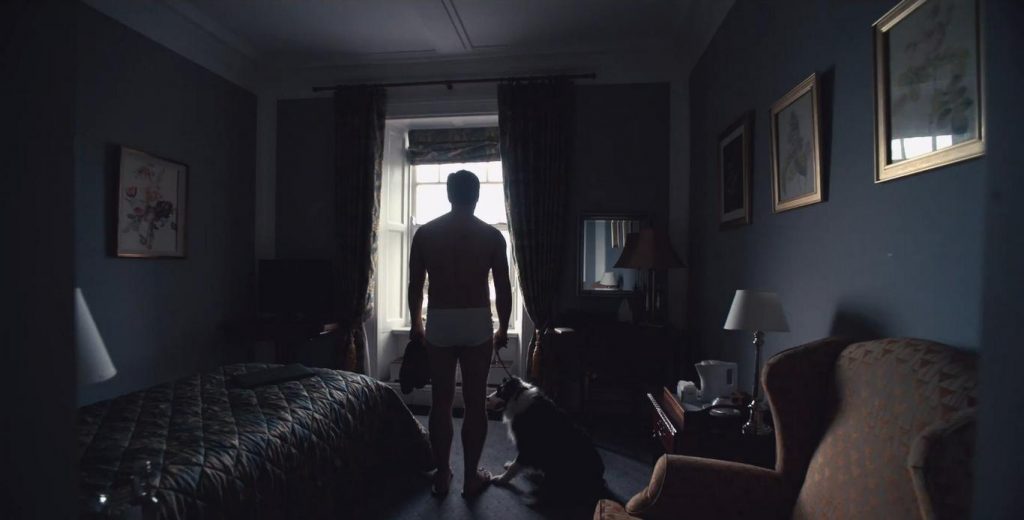 Hombre de espaldas en habitación oscura con cama y objetos personales, observando luz desde la puerta abierta