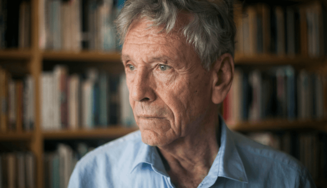Amos Oz, escritor israelí, en primer plano mirando hacia un lado con expresión pensativa frente a estantería de libros