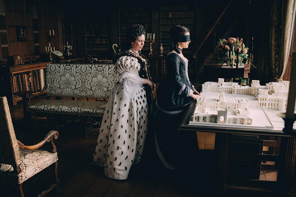 Dos mujeres en vestuario del siglo XVIII conversan en una sala opulenta con muebles antiguos y velas encendidas