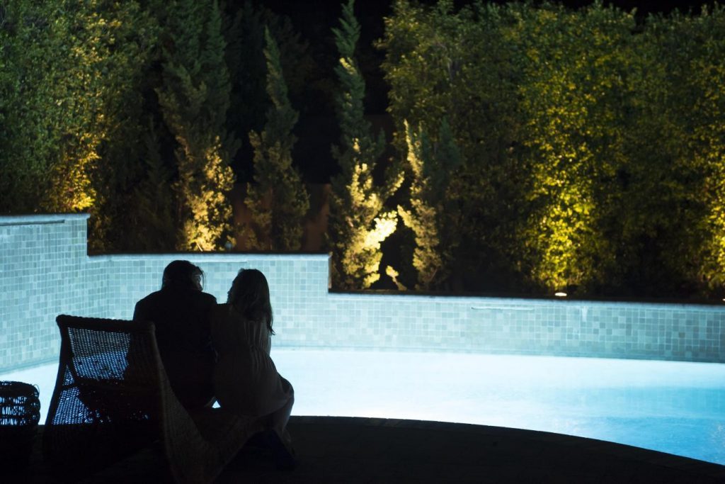 Dos siluetas frente a piscina iluminada de noche, árboles de fondo, ambiente festivo y tenso