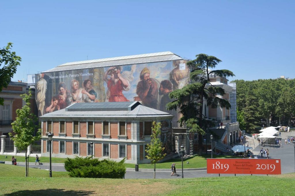 Fachada del Museo del Prado con gran mural de pinturas clásicas, banderas conmemorativas y jardines frondosos