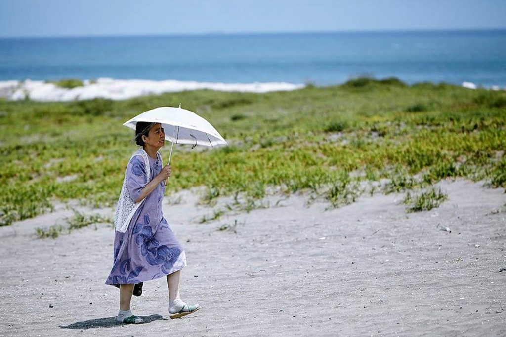 Mujer con sombrilla blanca caminando en playa arenosa con vegetación, cielo nublado al fondo