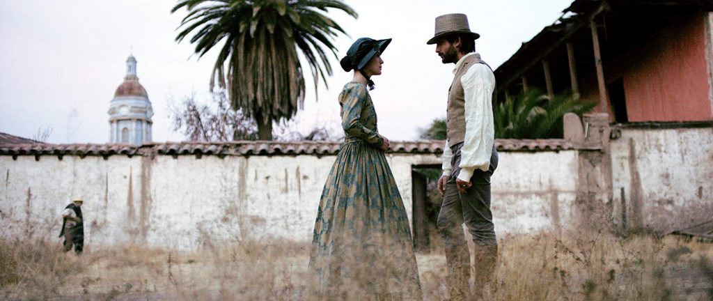 Pareja en traje antiguo frente a mausoleo colonial con palmera, escena de drama histórico chileno del siglo XIX