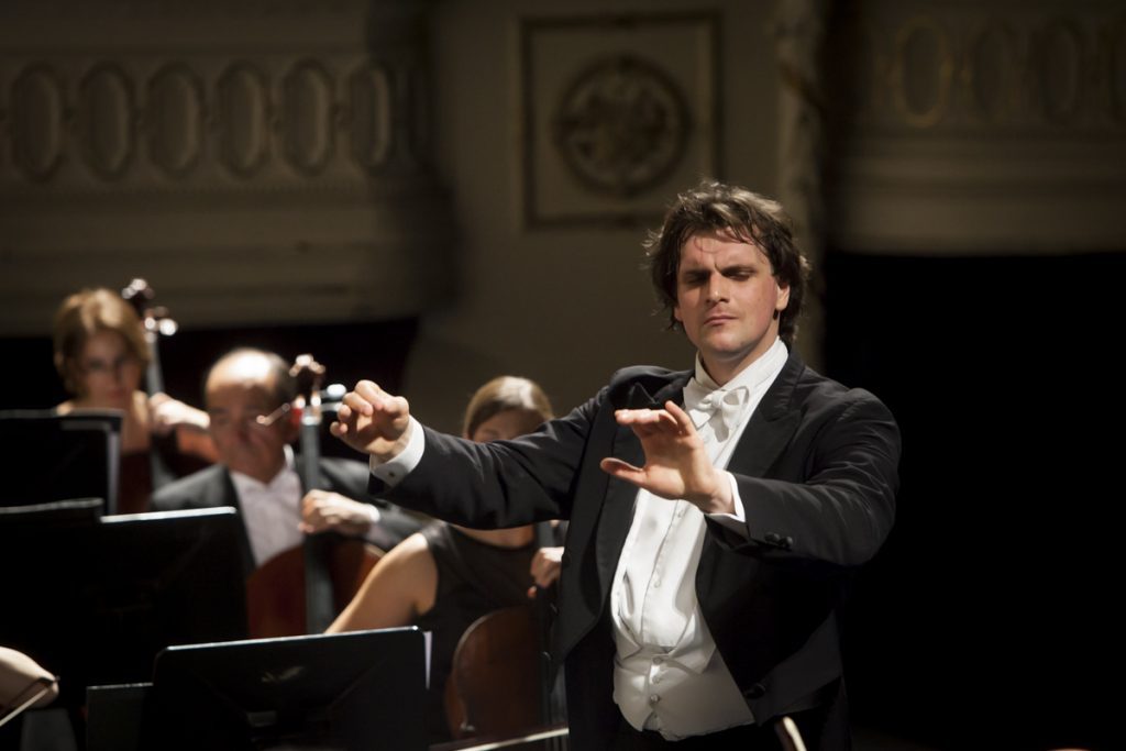 Director de orquesta con traje negro dirigiendo durante concierto en sala con músicos de fondo