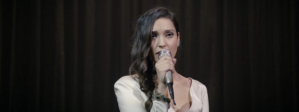 Mujer de cabello oscuro ondulado hablando al micrófono con fondo oscuro, durante presentación o conferencia