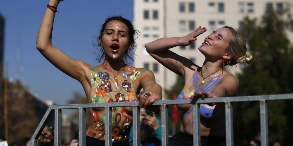Dos mujeres en una marcha feminista gesticulan desde un balcón con edificios al fondo bajo cielo despejado