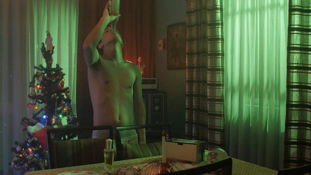 Escena interior nocturna: hombre sin camisa frente a espejo con árbol navideño de fondo y cortinas verdes iluminadas