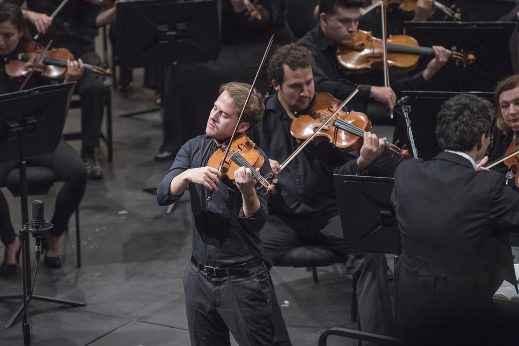 Violinista solista en primer plano tocando durante un concierto de orquesta, con músicos de fondo en escena