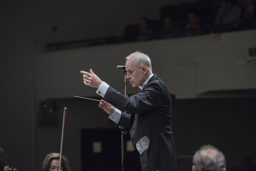 Concierto 3 del Ceac de la Universidad de Chile: Mosaicos imprevistos Conductor de orquesta con bastón en alto durante concierto, dirigiendo a músicos en escenario de sala de conciertos