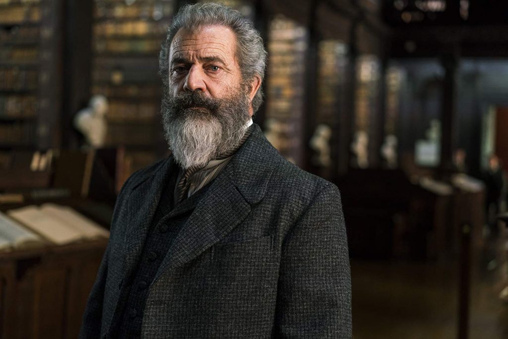 Hombre de mediana edad con barba gris y anteojos, vistiendo blazer gris oscuro en interior de biblioteca