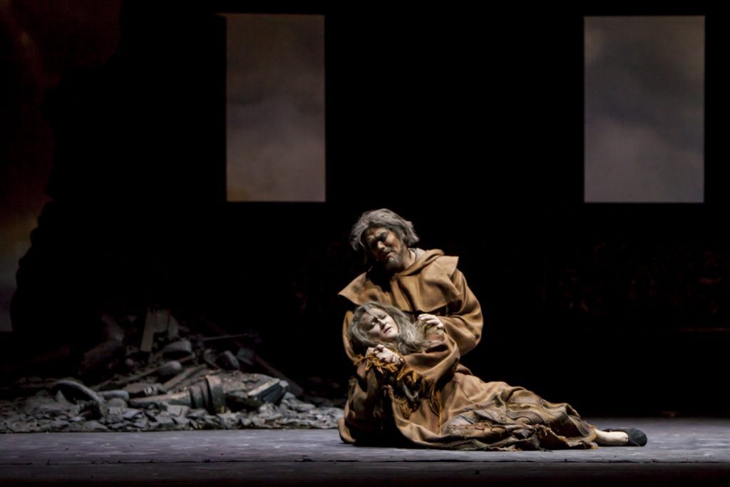 Actor en traje dorado arrodillado en escena teatral con ruinas y ventanas al fondo, representando momento dramático