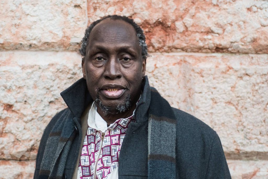 Ngugi wa Thiong'o, escritor keniano, retrato de frente vistiendo camisa cuadriculada y chaqueta oscura contra pared