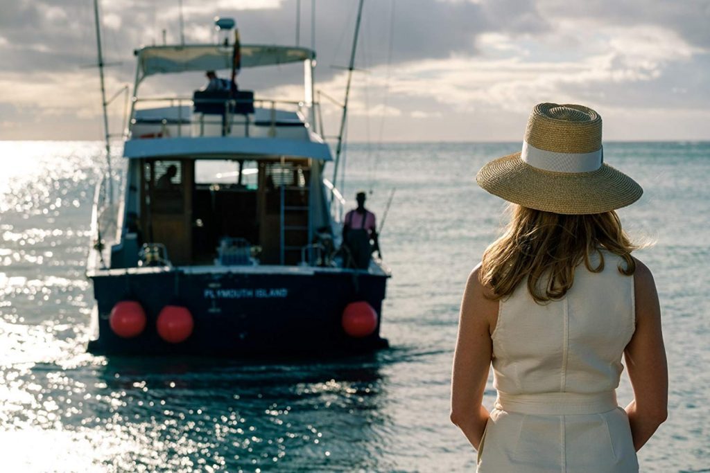 Mujer de espaldas en vestido blanco observa embarcación azul en aguas turquesas del Caribe bajo cielo nublado