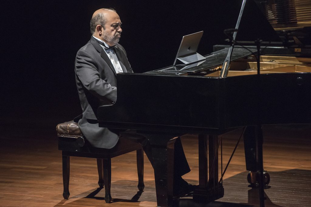 Pianista Alexandros Jusakos en concierto, tocando un piano de cola en escenario iluminado con fondo oscuro