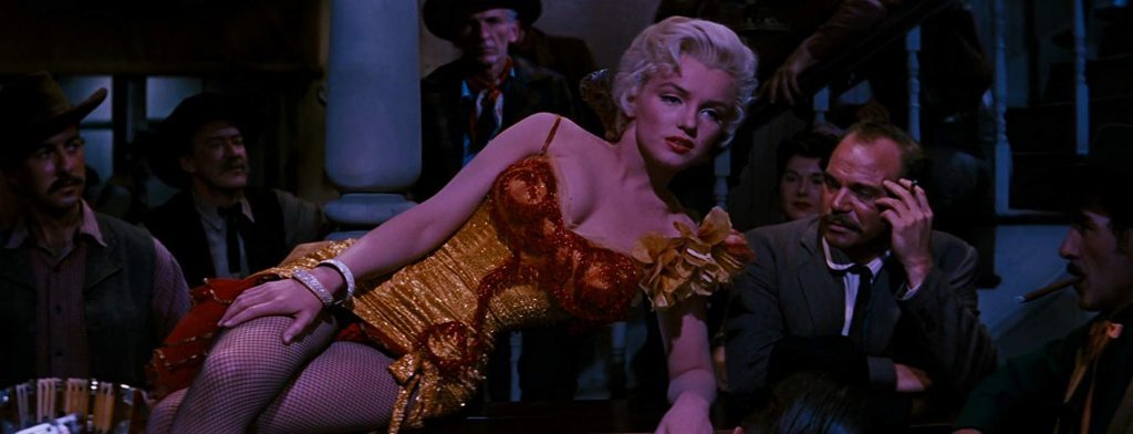 Marilyn Monroe recostada sobre una mesa en escena de cine, rodeada de hombres en traje, con iluminación azul