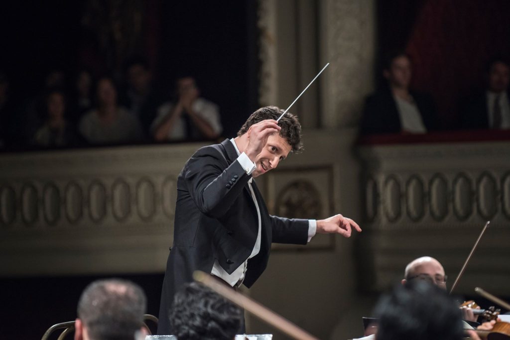 Conductor de orquesta dirige con batuta en un escenario de teatro, músicos de fondo y público en palcos
