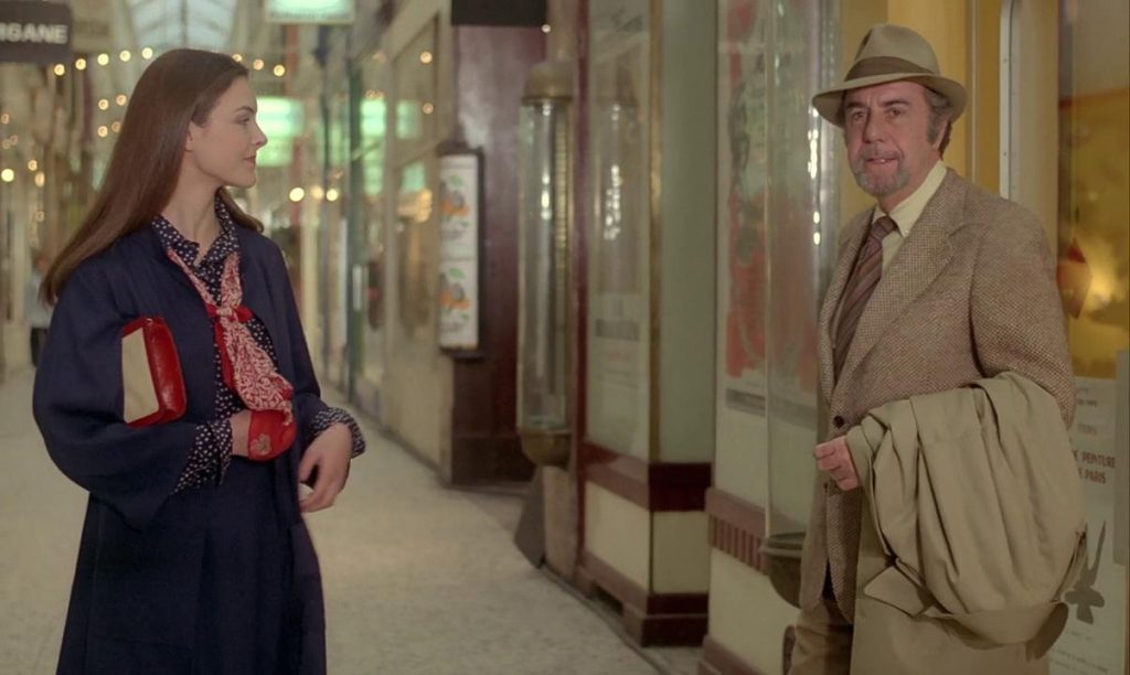Dos personas conversan en un pasillo elegante con vitrinas iluminadas de fondo, ella con abrigo azul y él con traje