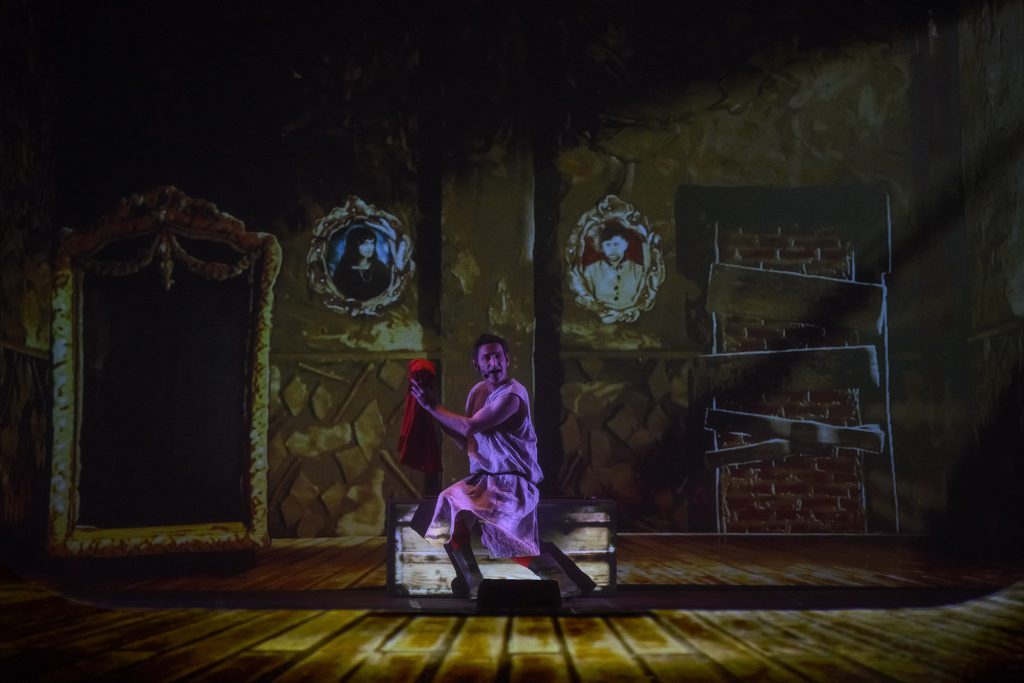 Escena teatral con figura en túnica púrpura frente a espejo ornamentado en decorado gótico oscuro y atmósfera lúgubre