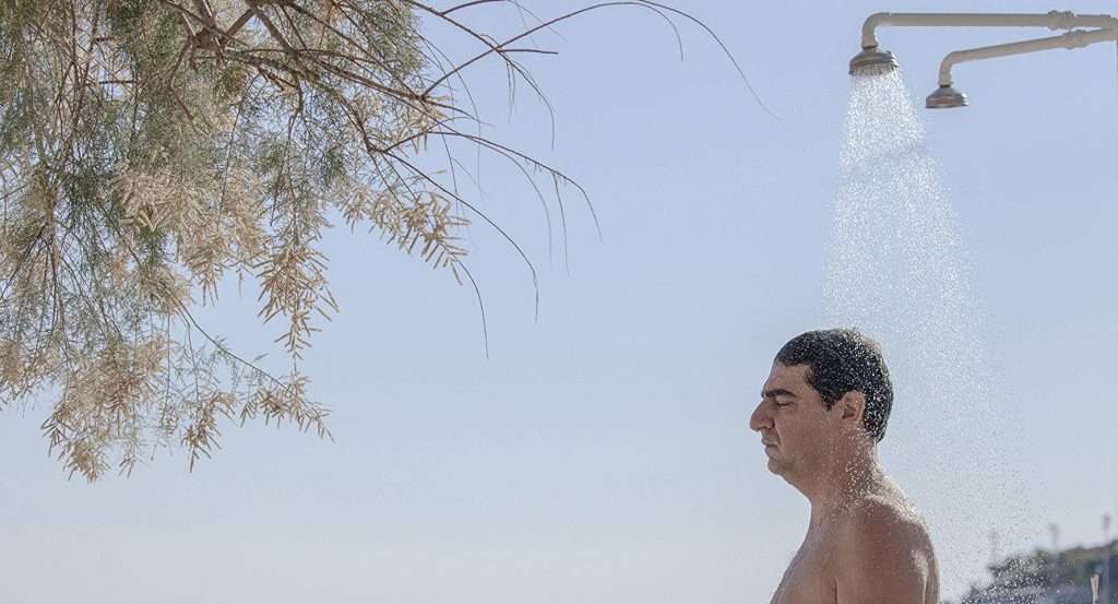 Hombre de perfil bajo una ducha, con árbol y cielo despejado de fondo, expresión contemplativa y melancólica