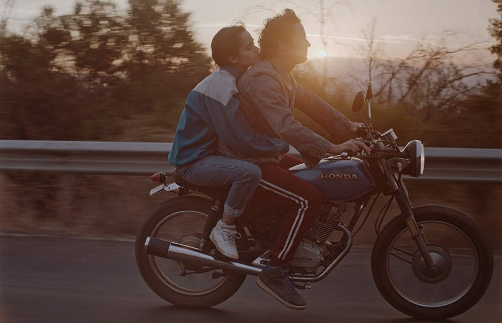 Dos jóvenes en motocicleta al atardecer, luz dorada, carretera y árboles de fondo, momento íntimo en movimiento