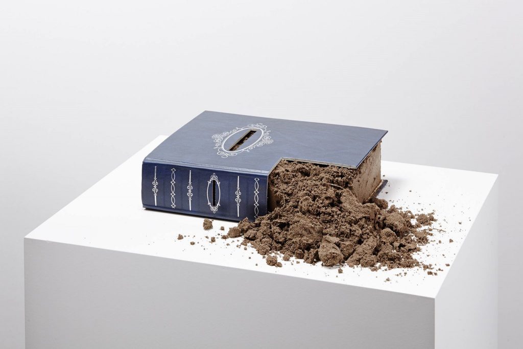 Libro azul oscuro junto a montículo de tierra marrón sobre base blanca, representando metáfora de lengua sepultada