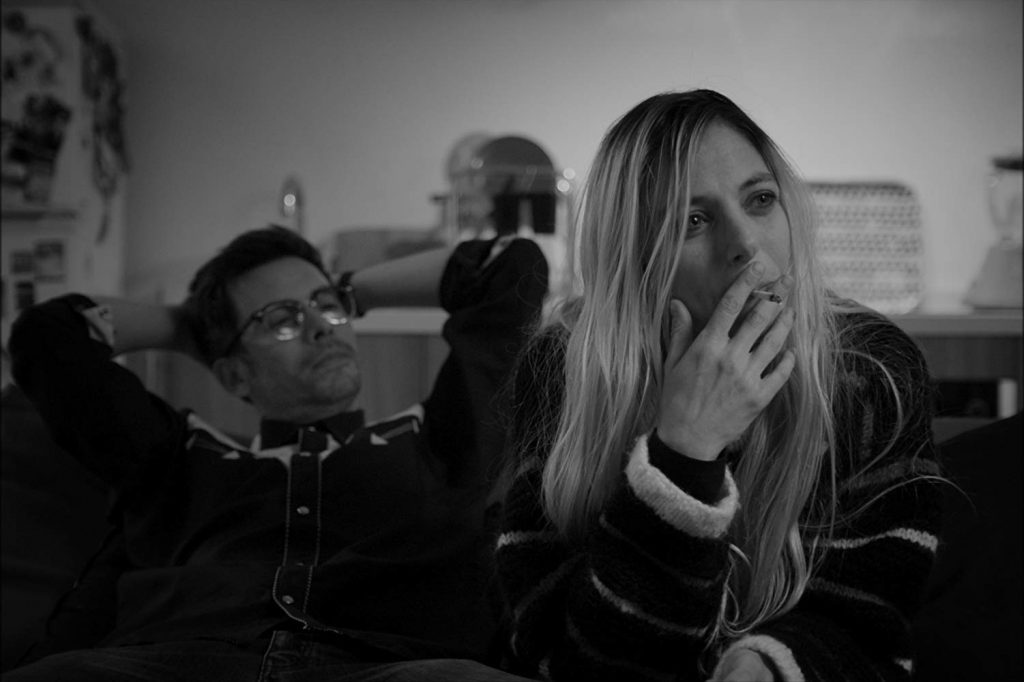 Mujer de cabello largo fumando en primer plano, hombre de fondo, interior en blanco y negro, atmósfera cinematográfica