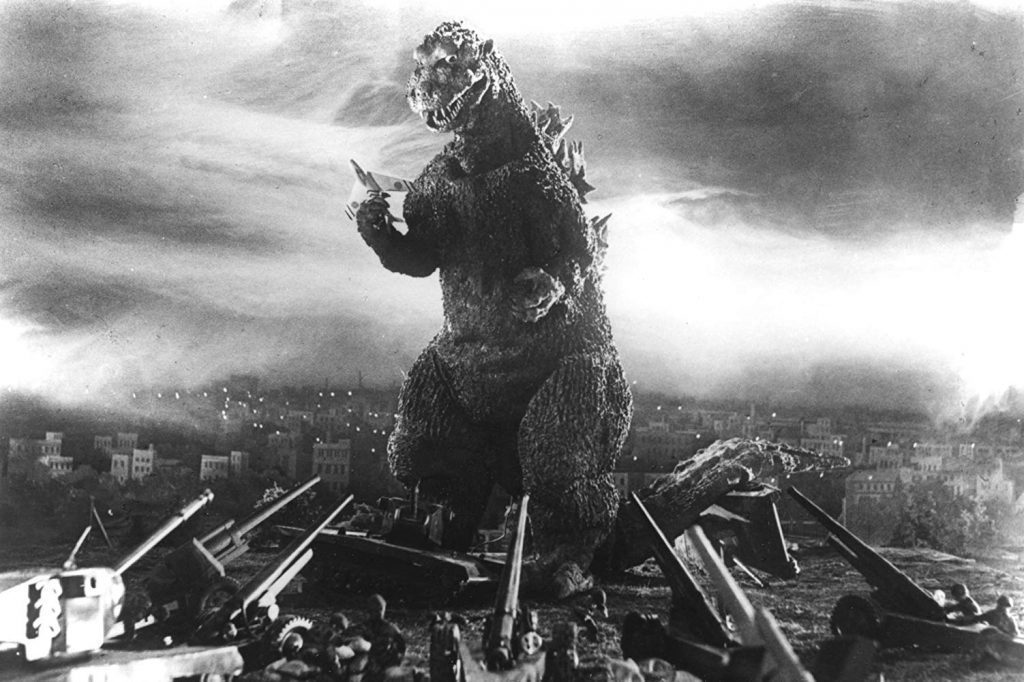 Godzilla emerge destructivo sobre ciudad en ruinas, respirando fuego, con edificios derribados y cielo nublado