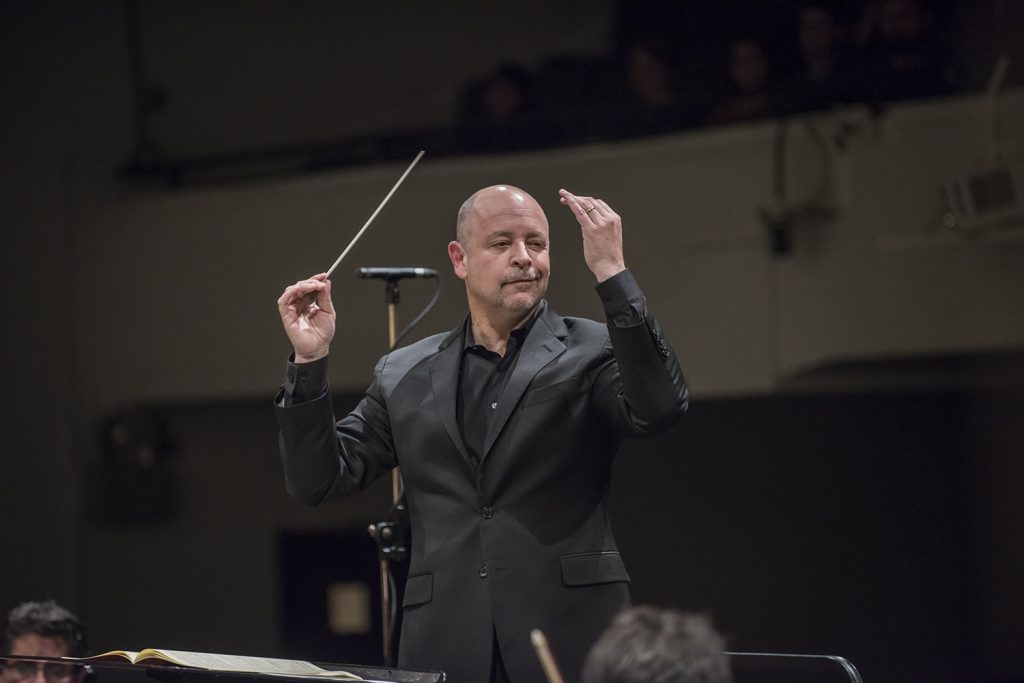 Director de orquesta José Luis Domínguez dirige con batuta durante concierto, vistiendo traje oscuro en escenario
