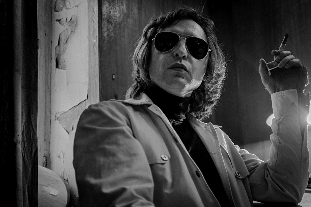 "Leto": La revolución silenciosa Hombre con gafas de sol y abrigo claro en blanco y negro, fumando en interior oscuro con pared desconchada