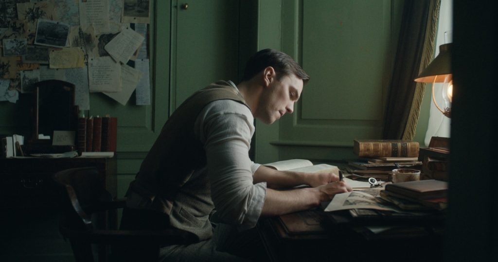 Hombre escribiendo en escritorio oscuro con lámpara, rodeado de papeles y libros en habitación con pared verde