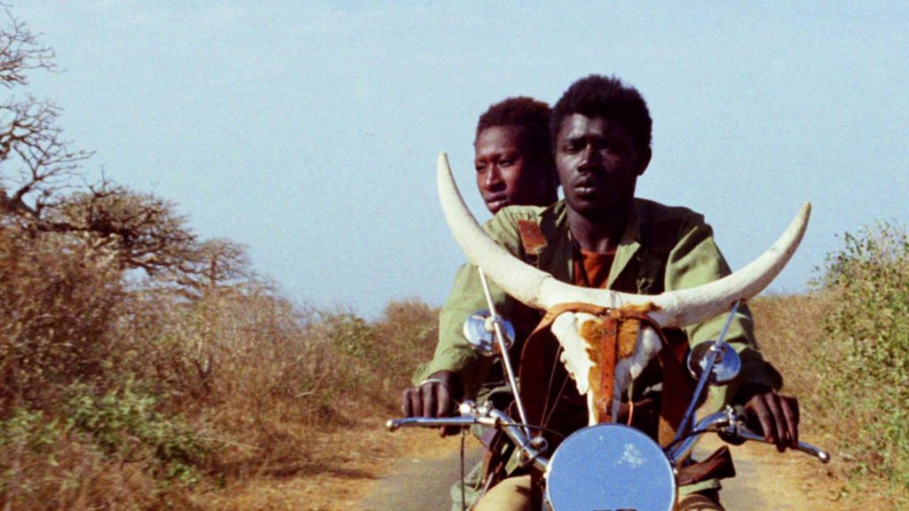 "Touki Bouki", de Djibril Diop Mambéty: El simbolismo de lo cierto Dos jóvenes en motocicleta con cuernos de buey decorativos, paisaje árido de sabana al fondo