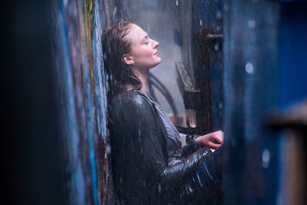 Mujer con cabello mojado bajo lluvia, apoyada contra pared azul oscura, expresión de intensidad y dolor emocional