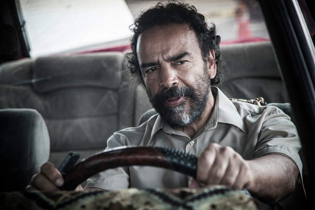 Hombre con barba al volante de un automóvil, mirando hacia adelante con expresión seria en escena de drama
