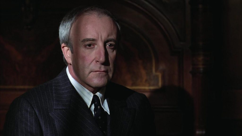 Peter Sellers con traje oscuro y corbata, en interior con iluminación cálida, expresión seria y contemplativa