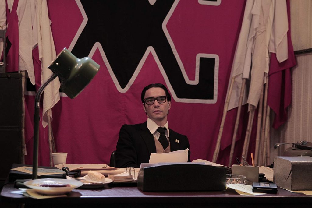 Hombre con gafas sentado a escritorio en oficina con pared magenta, frente a máquina de escribir y documentos