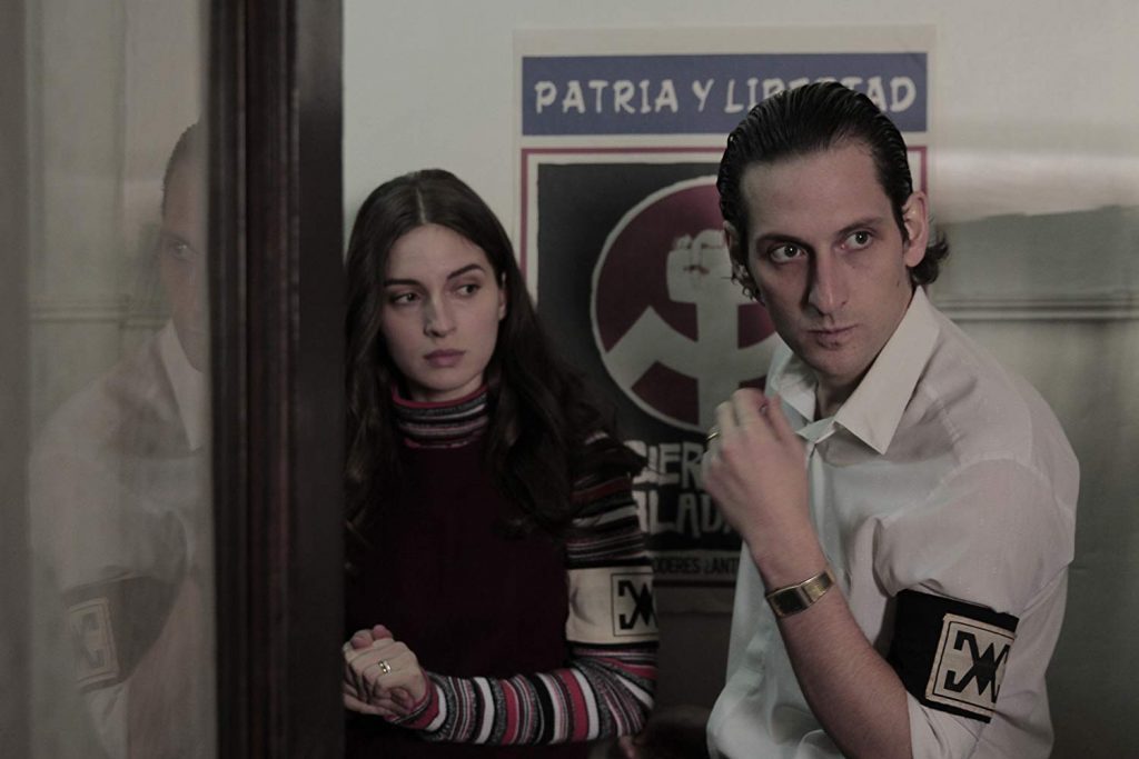 "Araña", de Andrés Wood: Los fantasmas errantes de Chile Dos personas en un interior oscuro junto a carteles de cine; mujer de cabello largo en suéter oscuro y hombre de camisa