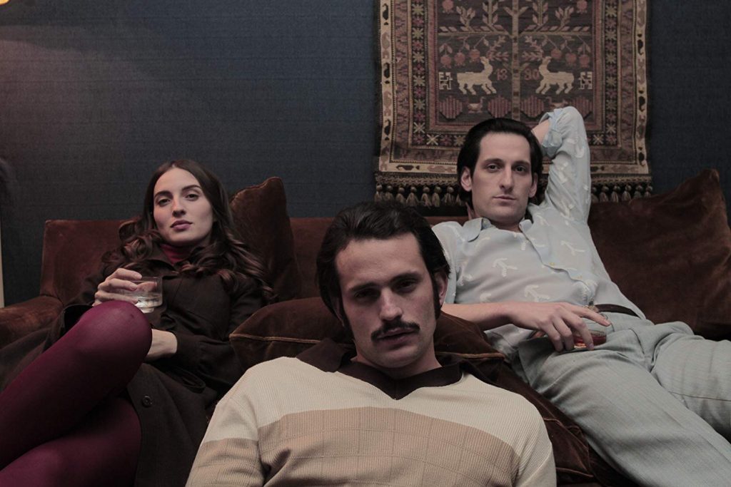 Tres hombres en interior oscuro reclinados en sofá burgundy, escena de la película Araña sobre conspiración política
