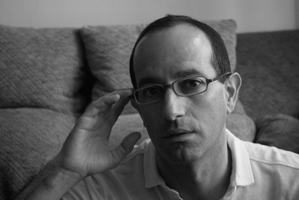 Nicolás Poblete Pardo, escritor, retratado en blanco y negro ajustándose las gafas frente a un sofá gris