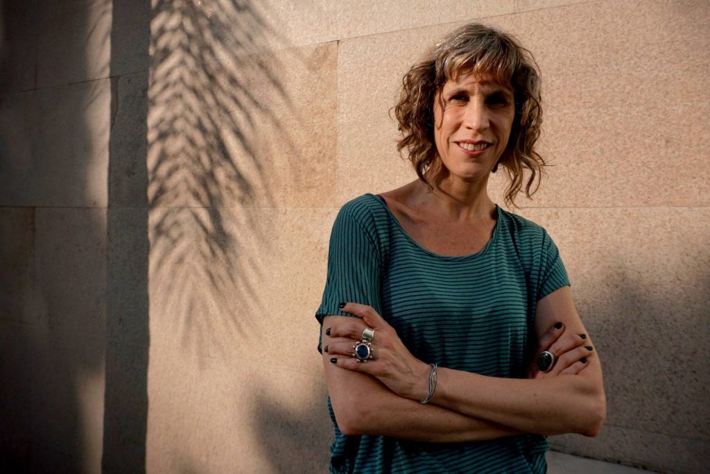 Nona Fernández, autora chilena, cruza brazos frente a pared beige con sombra de palmera, vistiendo camiseta azul verde