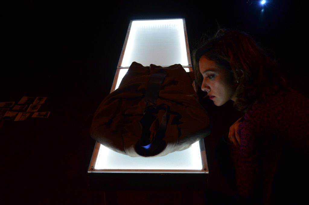 Persona sostiene un objeto sobre una caja de luz blanca en ambiente oscuro, evocando exploración visual y memoria