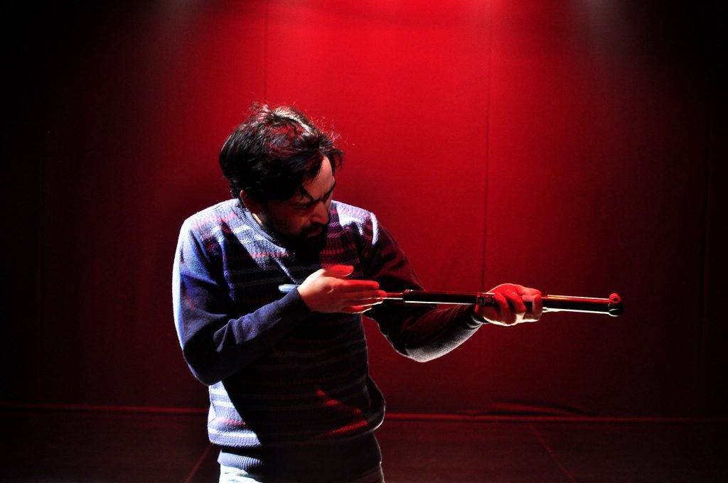 Actor en escena sostiene rifle durante representación teatral sobre memoria política, fondo rojo intenso