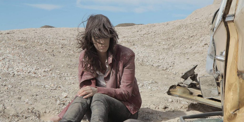 "Al desierto": Libertad condicional Mujer de cabello oscuro sentada en terreno árido y rocoso, vistiendo chaqueta marrón, mirando hacia abajo pensativa