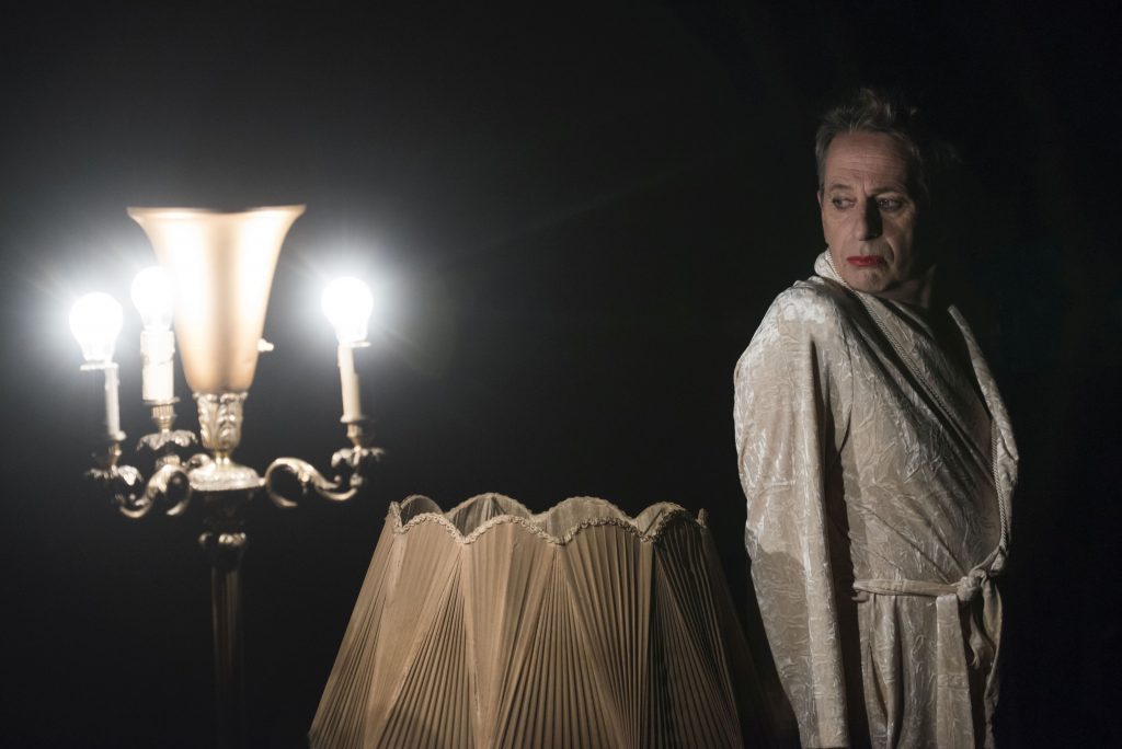 "Excesos": El palacio de la sabiduría Actor Alfredo Castro con túnica blanca ante lámpara encendida y cortinas de fondo, ambiente teatral