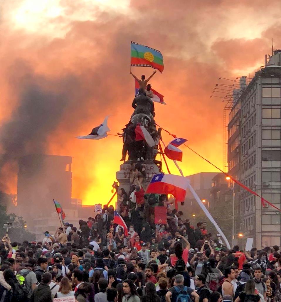 Multitud en plaza durante manifestación nocturna con edificios en llamas y bandera chilena en monumento improvisado