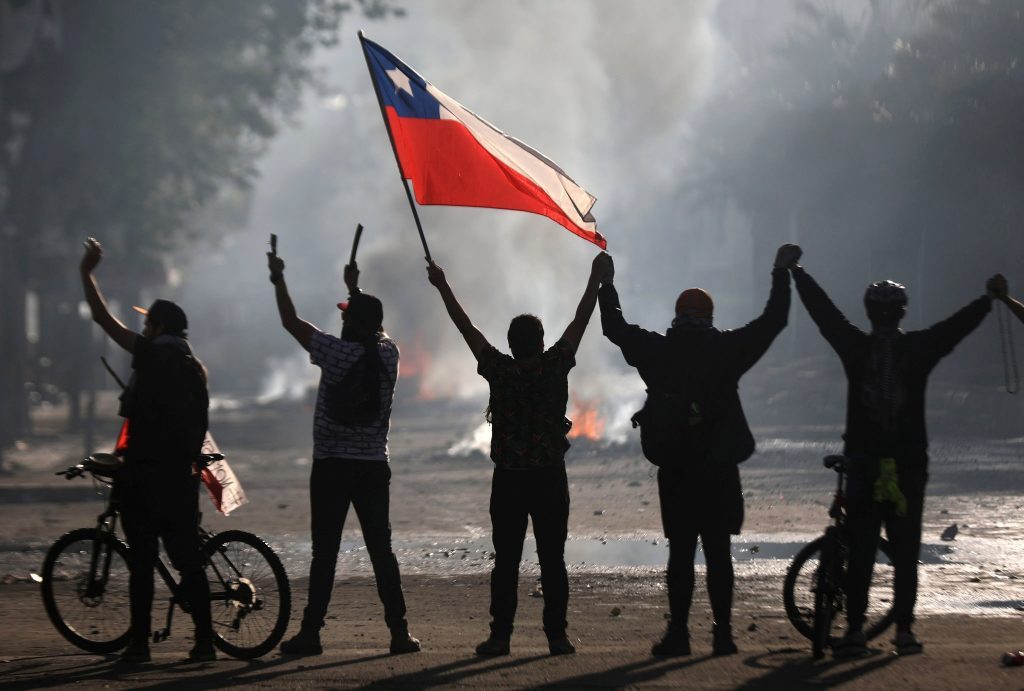 Cinco personas de pie con brazos levantados sosteniendo bandera chilena, con fuego y humo de fondo en protesta