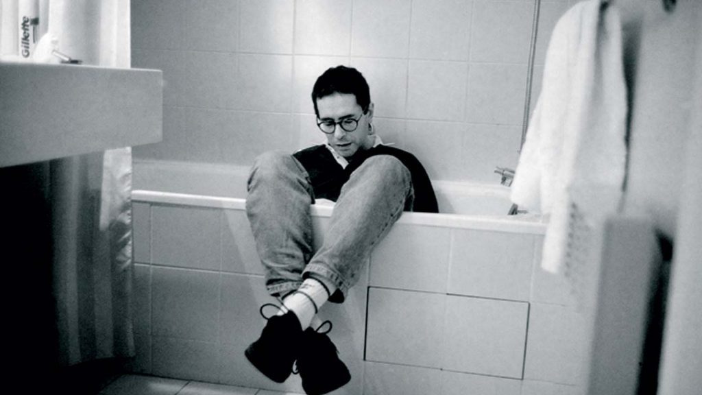 «El congreso de literatura», de César Aira: En el nombre del padre (Jorge Luis Borges) Hombre sentado en el piso de un baño blanco, abrazándose las rodillas, con expresión contemplativa y mirada hacia abajo