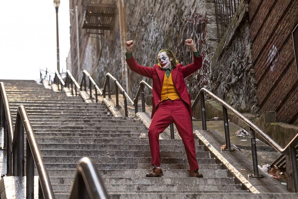 Hombre con traje rojo y amarillo en postura de brazos levantados, desciendo escaleras de piedra en zona urbana